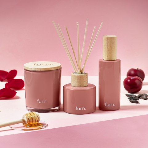 a gift set of home fragrance, including a candle, a room spray and a reed diffuser. the packaging is pink glass, and around them are ingredients of the scent, including sweet plums, honey and orange blossom.
