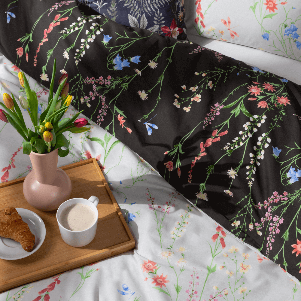 a breakfast in bed scene with a floral duvet cover, a wooden tray with a vase of tulips, a cup of coffee and a croissant. 