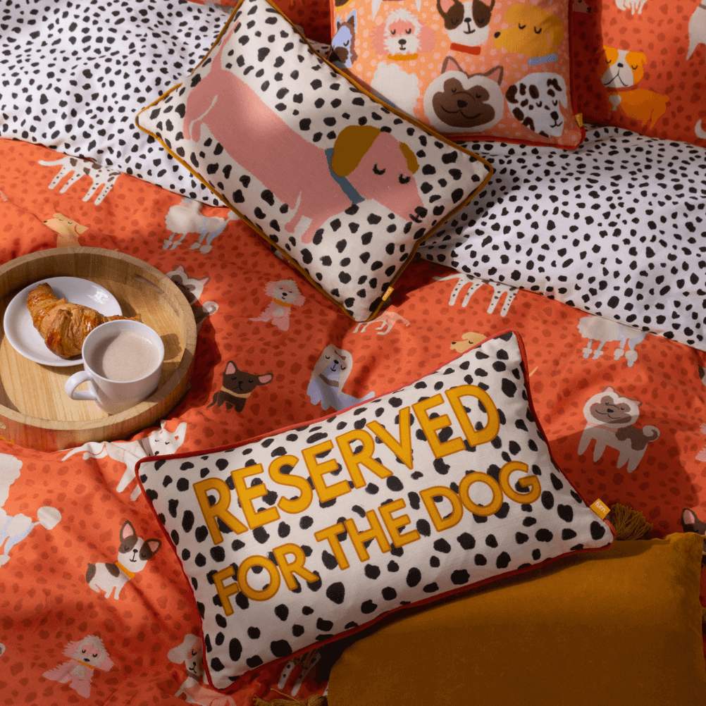 a breakfast in bed scene with the Woofers bedding and cushions, all featuring a dog themed print. there is also a round wooden tray with a cup of coffee and a croissant. 