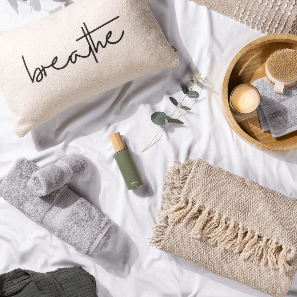 a self care flat lay, including a soft cushion with the word 'breathe' on it, a green scented room spray, a tasselled through and towels, as well as a tray with a lit candle and a body brush on it. 
