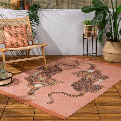 a wooden decking surface with a coral outdoor rug on it, with a tibetan tiger illustrated design, and a decking chair with a matching coral cushion on it.