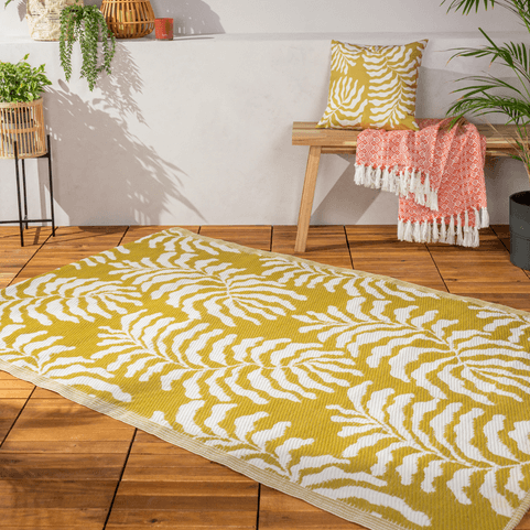 a wooden decking with a mustard coloured outdoor rug on it, with a soft white floral design, and a matching cushion sitting on a wooden deck chair in the background. 