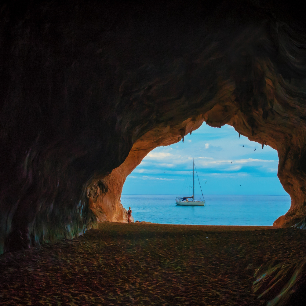 a large stone cave which opens out onto a bright blue sea, in which there is a small boat.