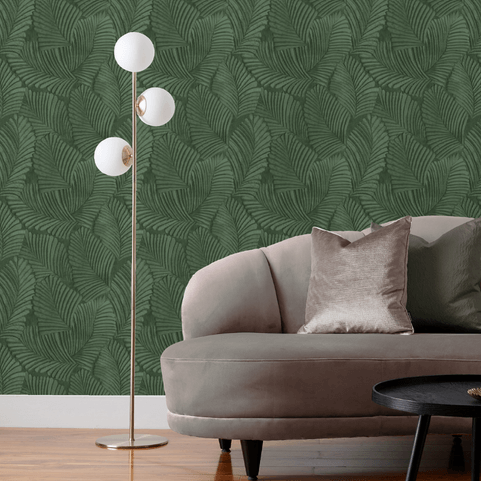 a wall with emerald green vinyl floral printed wallpaper, behind a three-bulb floor lamp and a grey velvet sofa with a matching cushion.