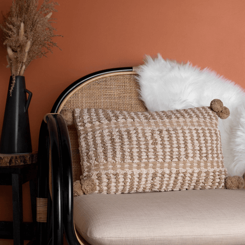 a natural-coloured woven jacquard cushion with pom pom detailing, on a beige chair with a white fur throw.