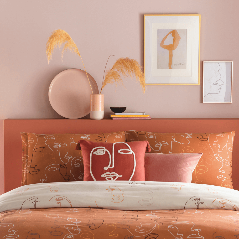 a pale pink bedroom with orange abstract bedding, with picture on the wall and a few decorative ornaments.