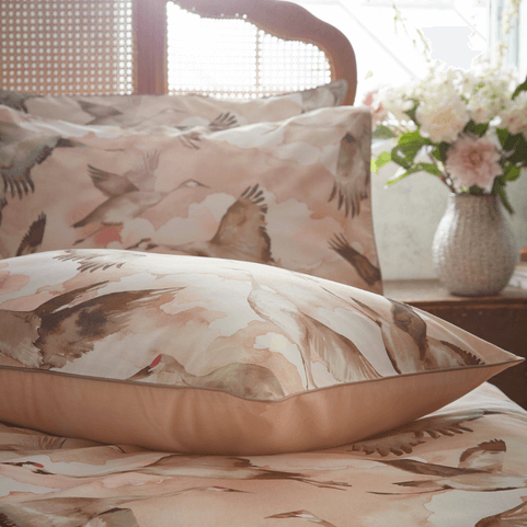 a bed with a blush pink patterned bedding on it. the bedding has a flock of storks in flight through the clouds on it. there's also a rattan headboard and a vase of pink and white flowers on the bedside table.