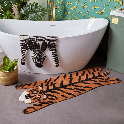 a tiger print and tiger shaped bath mat laid out on a grey bathroom floor, with a matching zebra bath mat draped over the side of a bath in the background.