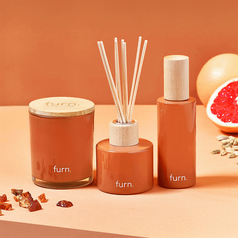 a collection of burnt orange home fragrance items placed in a row, including a glass candle, a reed diffuser and a bottle of room spray.