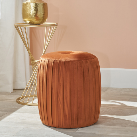 a luxury velvet pouffe in a tobacco orange shade, sitting on a grey floor with a gold side table in the background.