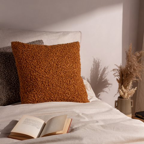 a ginger coloured boucle cushion with a matching grey cushion, propped up on a white bed.