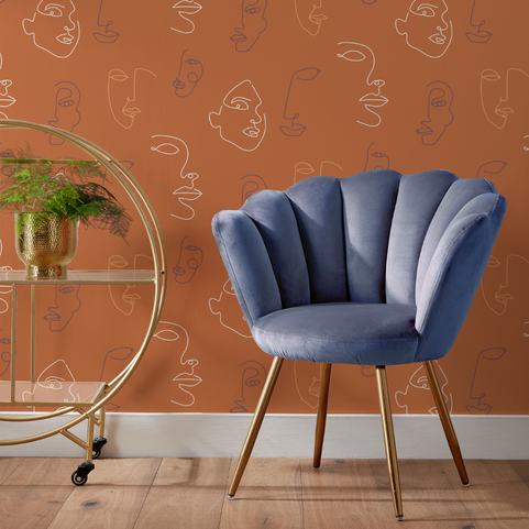 a terracotta orange wallpaper with an abstract face design, with a chair and wheeled gold shelving unit in the foreground.