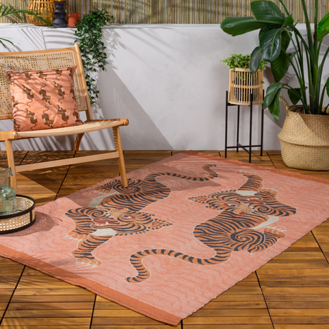 a coral orange area rug with a printed design of two tibetan tigers, laying on a wooden decking next to a rocking chair with a matching orange cushion on it.