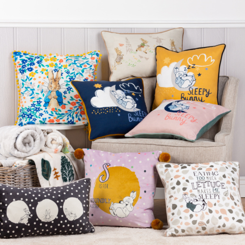 a collection of eight Peter Rabbit cushions arranged on a white side table, a beige armchair and a cream carpeted floor.