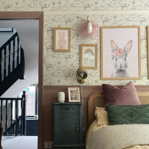 a bedroom with a muted pastel colour scheme, framed wall art of rabbits and a rainbow, a bed dressed with a muted purple and forest green geometric cushion, and neutral winter forest wallpaper.