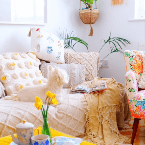 a neutral white living room decorated with a tufted sofa throw, tufted and textured white and beige cushions, a yellow tasseled throw, and a woven hanging flower pot. There is a pink floral chair on the right hand side, and a tea set with a daffodil vase in the foreground.