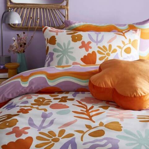 A multicoloured duvet cover set with an abstract floral design, made on a bed with a coordinating orange cushion, and folded to reveal the squiggly line reverse design.