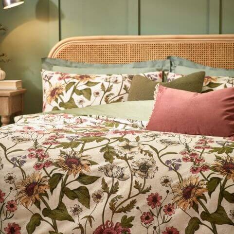 A closeup of a cream coloured duvet set with a multicoloured floral design, made on a bed with green and pink scatter cushions, a wooden headboard and a green wall in the background.