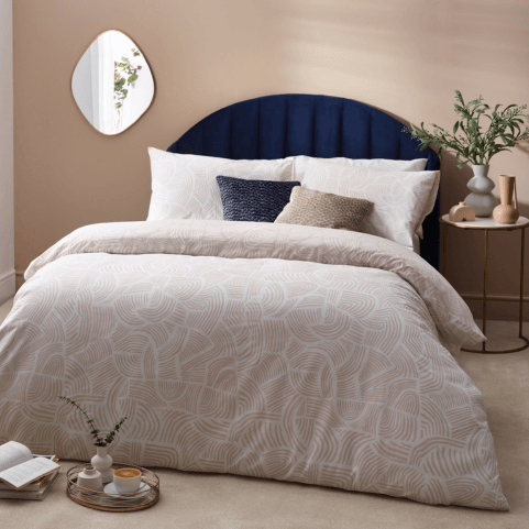 A swirling sand dune abstract patterned duvet cover set in a beige shell shade, dressed on a bed with coordinating beige and deep blue cushions. There is a side table with a potted plant and various ornaments next to the bed, and a series of ornaments lying on the floor in the foreground.