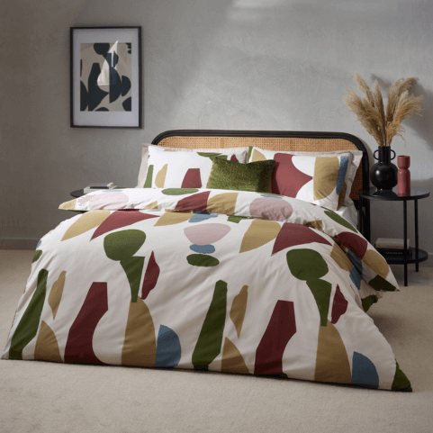 meta geometric abstract bedding dressed on a bed with a coordinating olive green velvet cushion. There is a side table with a potted plant and vase to the right, and framed wall art to the left.