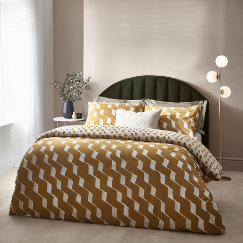 honey yellow bedding with an abstract crimped line pattern in white, dressed on a bed with a matching off-white velvet cushion. There is a floor lamp to the right and a potted plant and ceramic ornaments on a side table to the left.