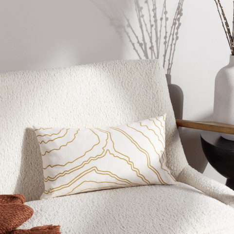 a nougat beige cushion with an abstract wavy line pattern in gold, sitting on a textured beige armchair.
