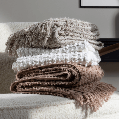 three woven bouclé yarn throws in feather, natural and plaster shades, piled on top of each other on a beige textured sofa.