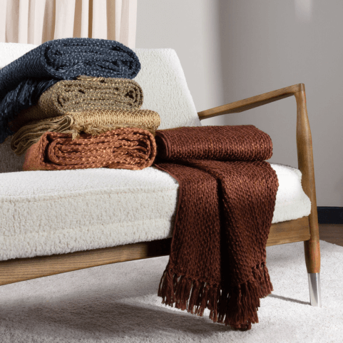 four woven yarn throws in dusk, olive, hazel and chestnut shades on an off-white textured sofa. Three are folded up in a pile, and one is folded and draped off the front of the sofa.