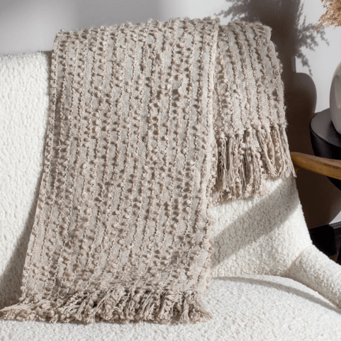 a neutral feather coloured woven bouclé yarn throw with soft textural fringing on two edges, folded and draped over the back of a white textured sofa.