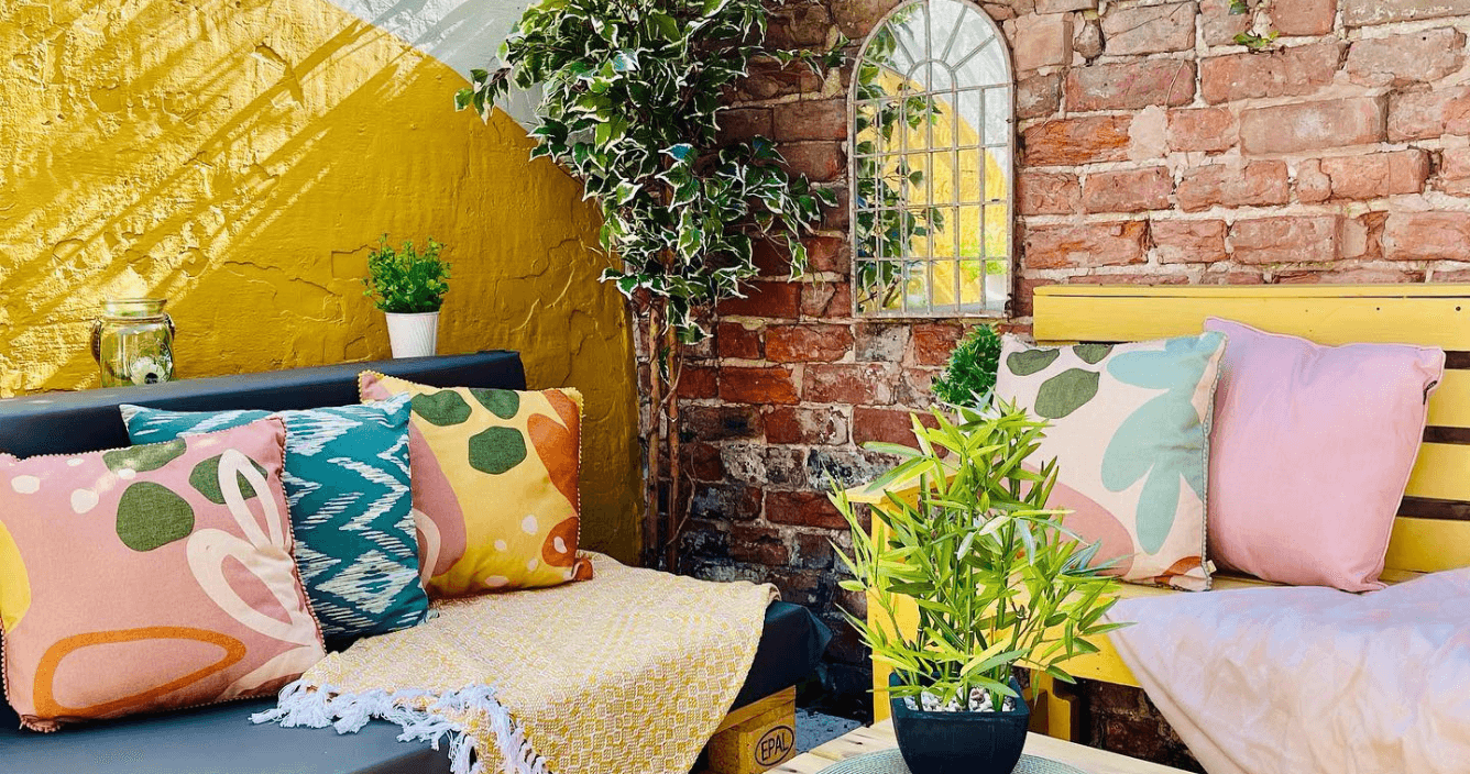 an outdoor garden space with seating, bright abstract and floral cushions, potted plants, and a yellow tasseled throw.