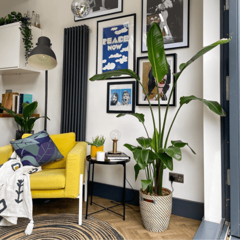 a living area with a seven-piece gallery wall, a yellow chair with a recycled blue-green leaf cushion and a tasseled boho throw on it, a potted plant, a side table with ornaments on it, and a large floor lamp in the background.