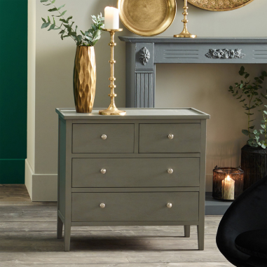 A wooden 4-drawer storage unit in a grey painted shade, standing in a neutral room holding a gold candelabra and a gold vase.