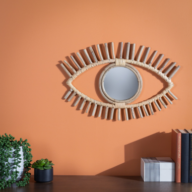 A woven rattan mirror designed in the shape of an eye, presented on an orange wall behind potted plants and books.