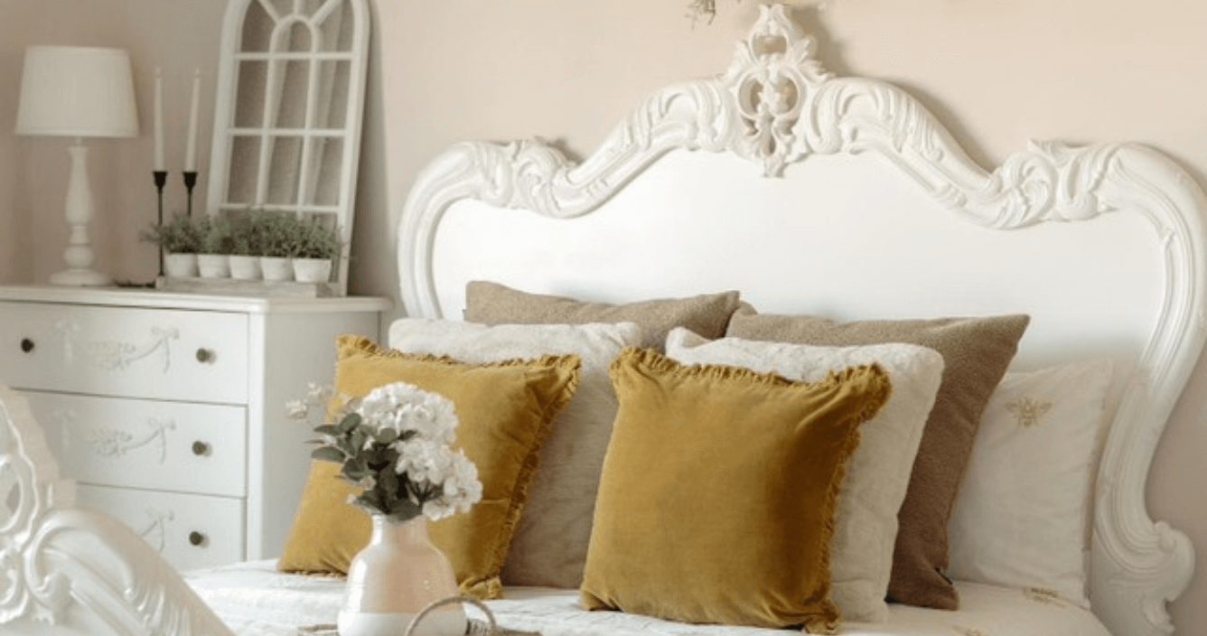 A royalcore style bedroom, with a white ornate French-style bedframe, lots of plush cushions and a vase of white flowers.