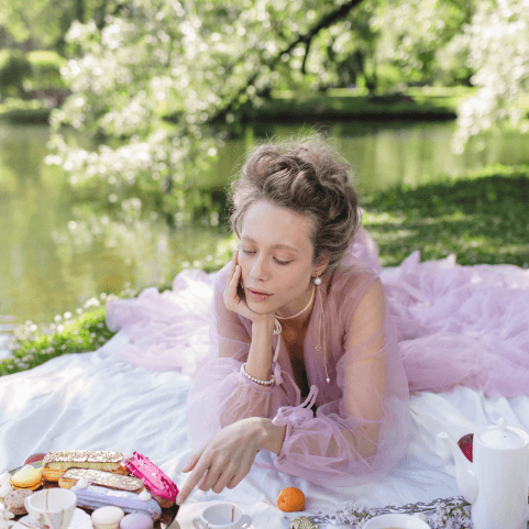 a woman in a pink tulle gown, hair piled on top of her head and pearl jewellery lies propped on her elbows on a white picnic blanket as she chooses which exquisite pastry she might eat next. 