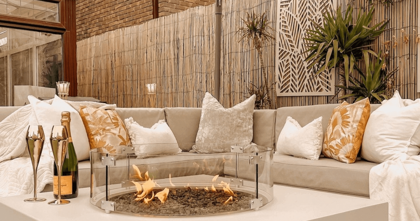 a grey outdoor sofa with an arrangement of white, grey, gold and fringed cushions, and two white sofa throws. There is a built-in fireplace table in the foreground with a bottle of champagne and two glasses, and there is a bamboo fence and trees in the background.