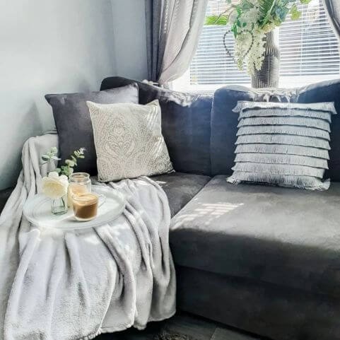 a grey corner sofa layered with an array of grey cushions, as well as a silver throw. there is a tray with flowers and a coffee on.
