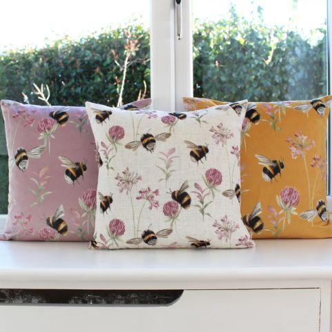A set of three of the same cushion in white, lilac and gold, respectively. The pattern features bees and delicate flowers.