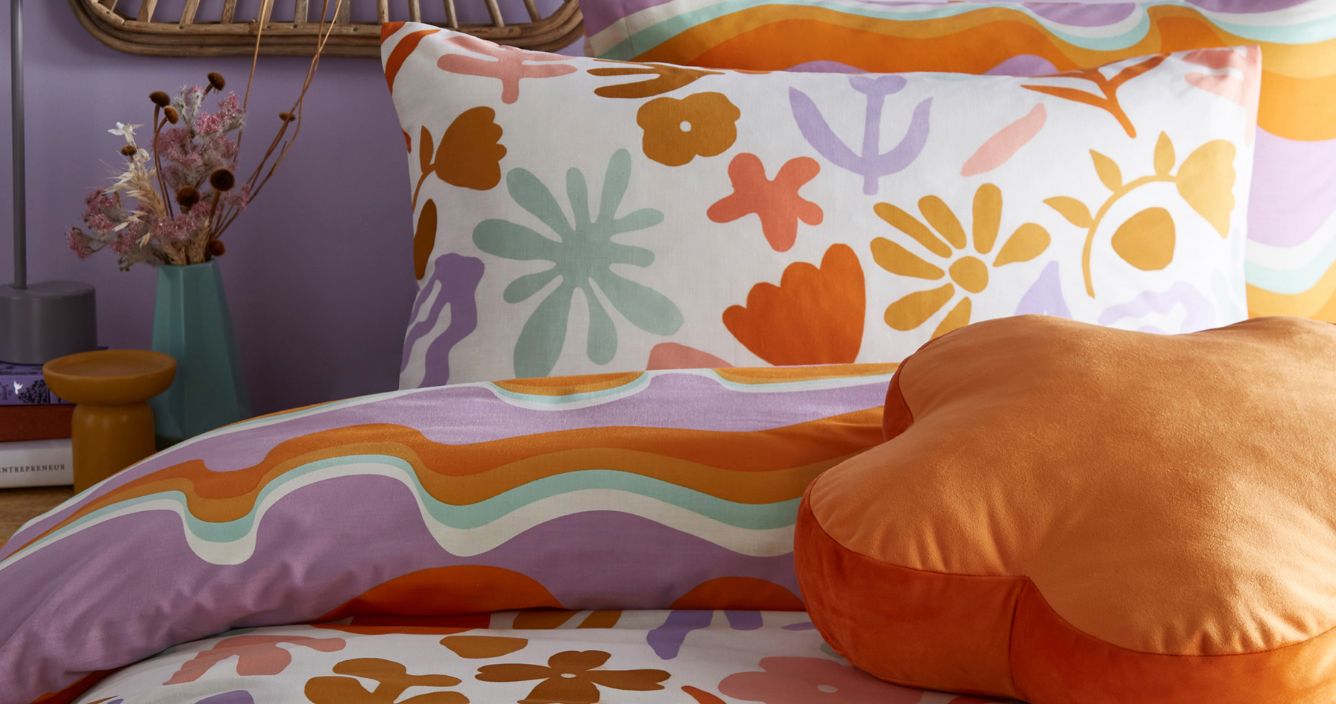 A graphic floral bedding set in orange, lilac, pale green and yellow on a bed. There's an orange flower-shaped cushion resting on top.
