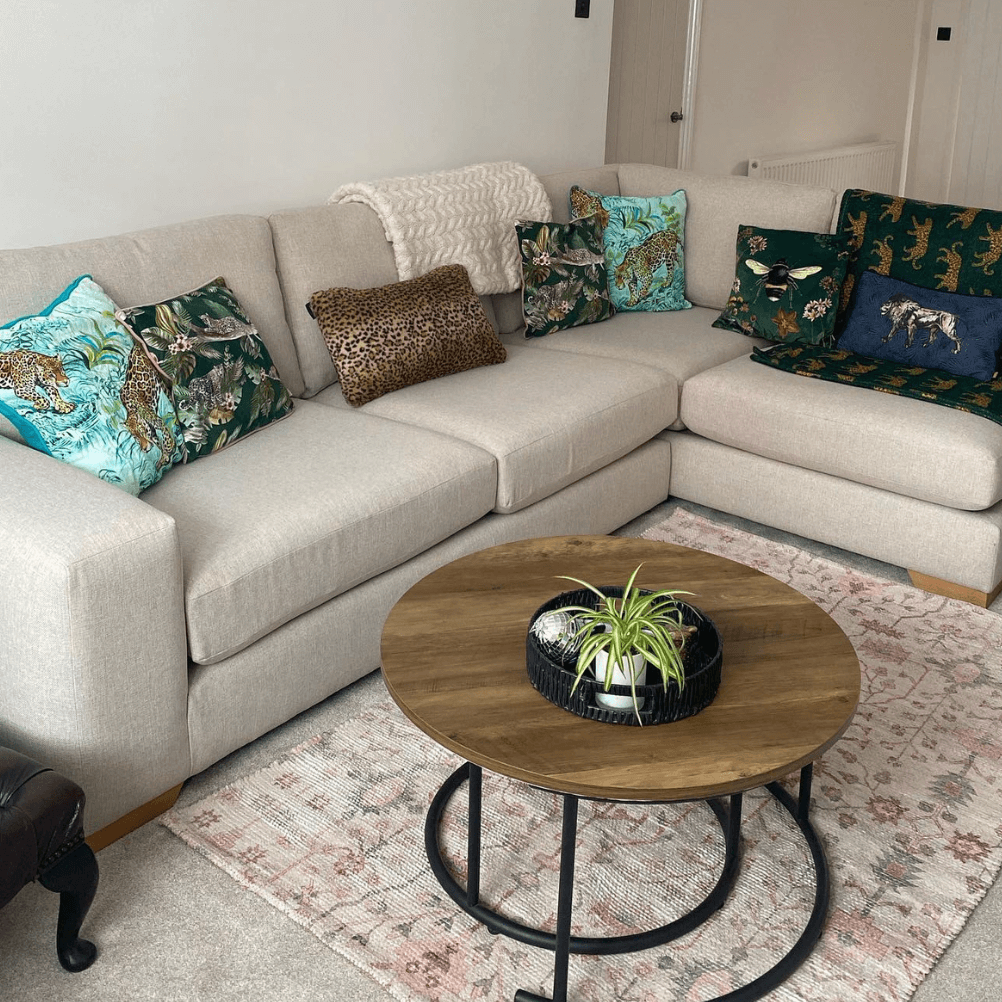 A neutral living room with a round wooden coffee table, a woven rug and a grey sofa decorated with scatter cushions sporting animal, exotic and animal print designs.