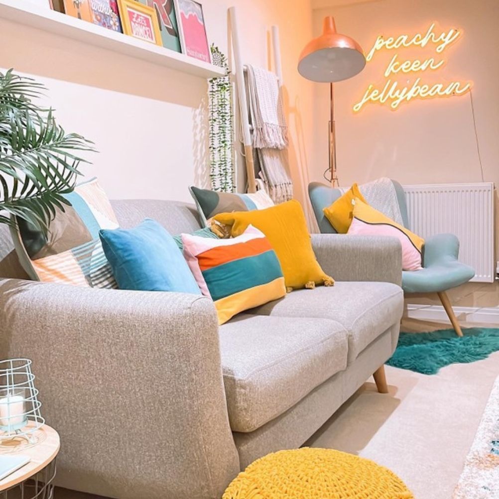 A bright living room with a neon wall sign, large floor lamp, a display shelf and indoor plants, with a grey sofa holding a selection of cushions in plain, tasseled and geometric designs.