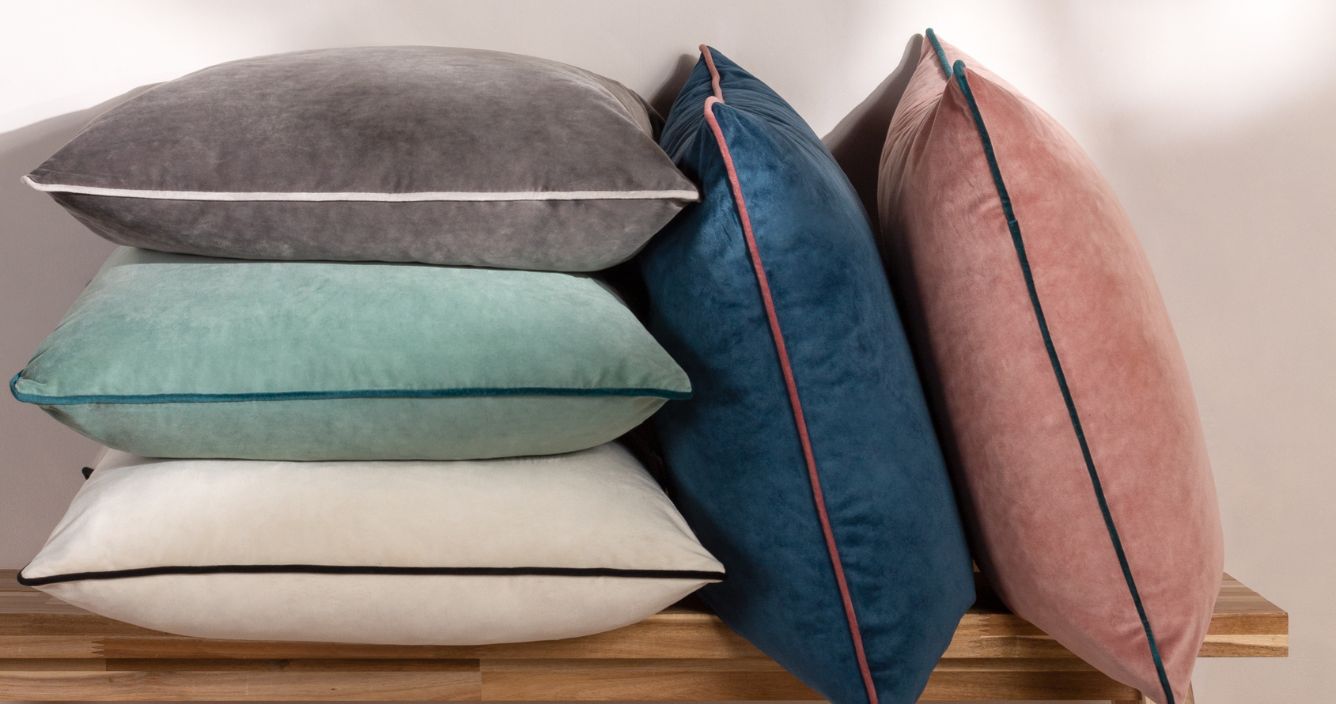 A pile of velvet cushions of all different colourways, three cushions are piled on each other and two are leaning. 