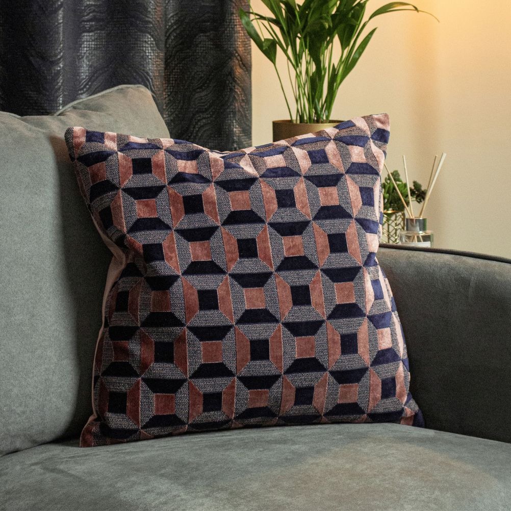 A pink and blue velvet geometric pattern cushion on a grey sofa. There is soft light, a plant and a curtain in the background.