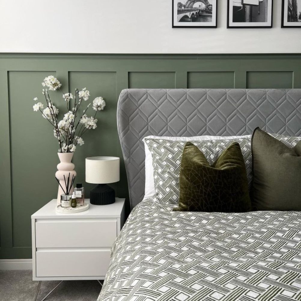 A bed with two green cushions on a green and white duvet cover set. There is green panelling, and cream walls, there's a bedside table with a bunch of white flowers in a vase.