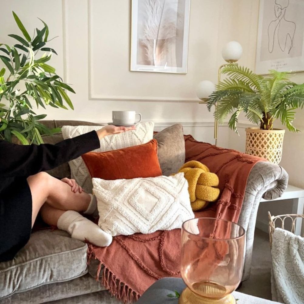 A grey velvet sofa piled high with different colour and size cushions. There's a person holding a cup up over the cushions.