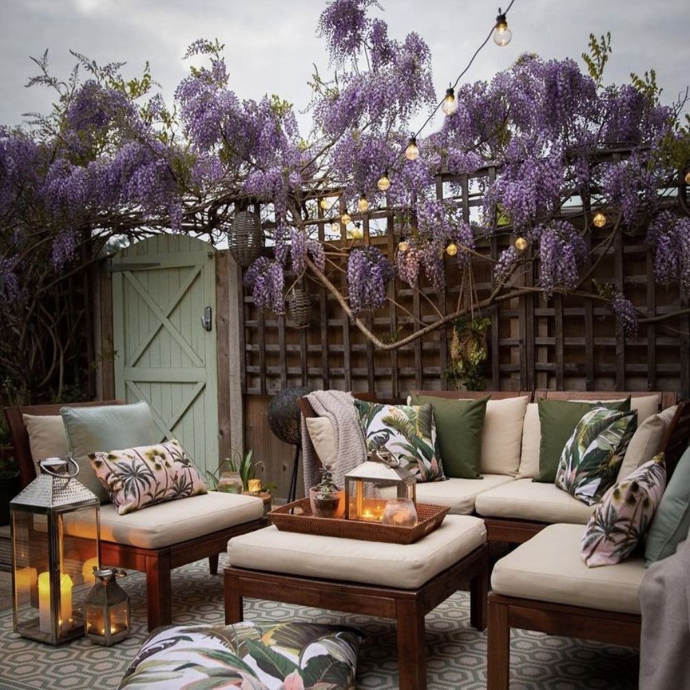 An outdoor patio scene with purple wisteria hanging above a set of wooden outdoor furniture. It's decorated with plain green and leafy patterned outdoor cushions.
