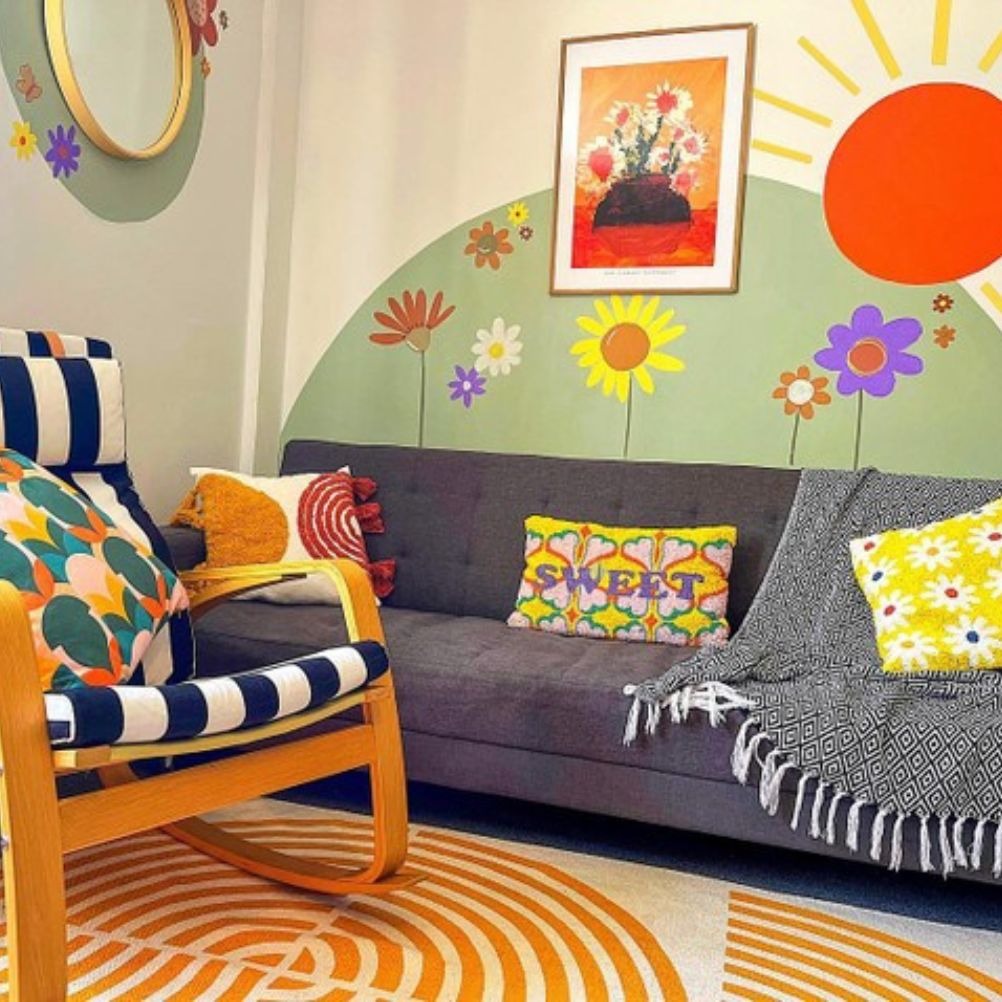 a maximalist style living room, with graphic flowers painted on to the wall. The sofa is grey with bright knitted cushions. 