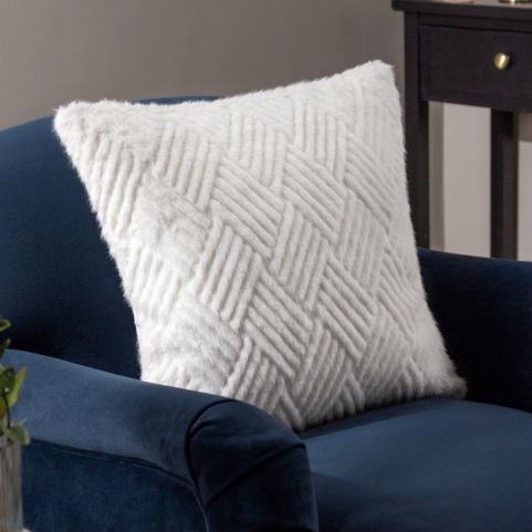 A white faux fur cushion with a cut geometric design, displayed on a navy blue sofa.