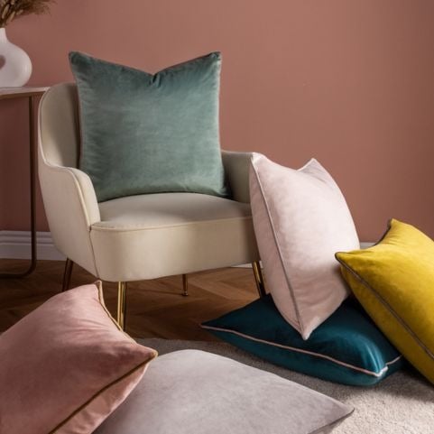 A selection of velvet cushions in a variety of muted and bright shades, arranged on a cream coloured sofa chair.