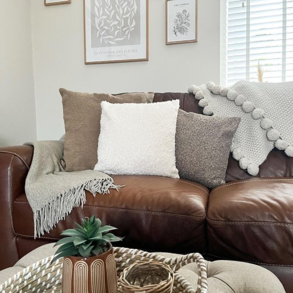 A brown leather 2-seater sofa with three cushions on one side, with a couple of throws thrown over it. 
